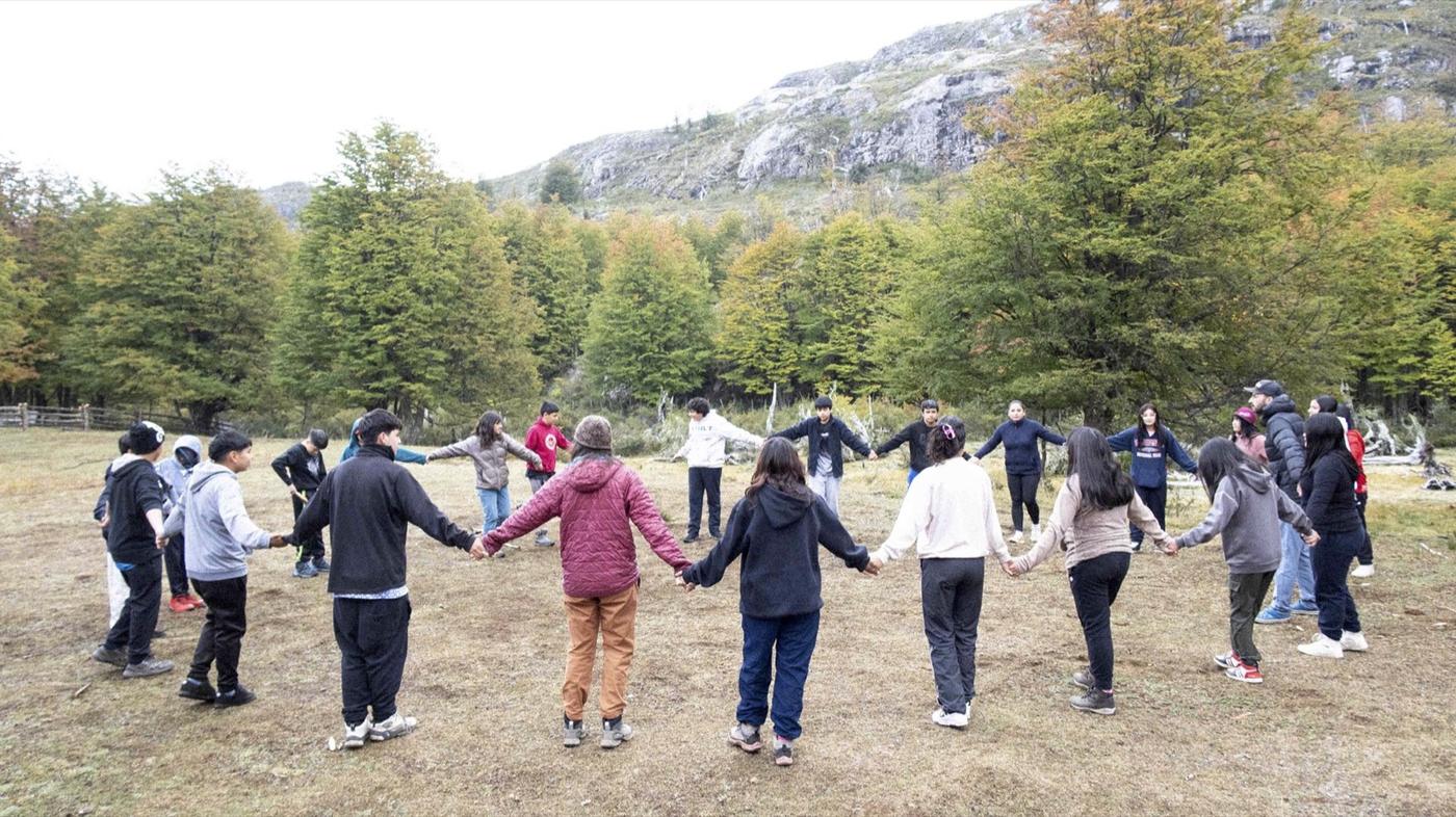 Un grupo grande de estudiantes se toma de la mano en ronda sobre el pasto, frente a los cerros del Parque Escuela Kaikén